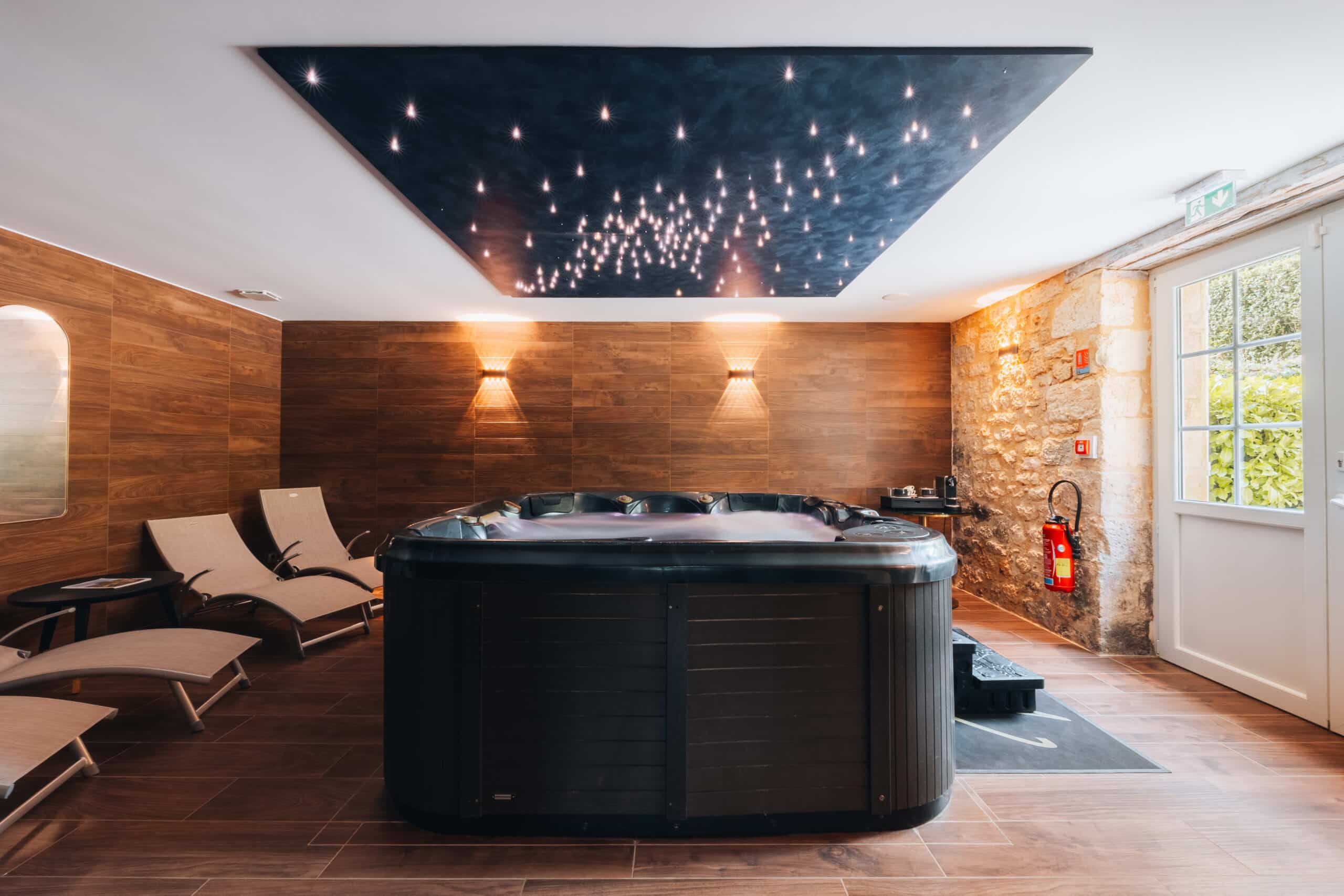 Espace spa intérieur avec jacuzzi et transats dans une ambiance chaleureuse, bois et pierre, éclairage tamisé et ciel étoilé au plafond